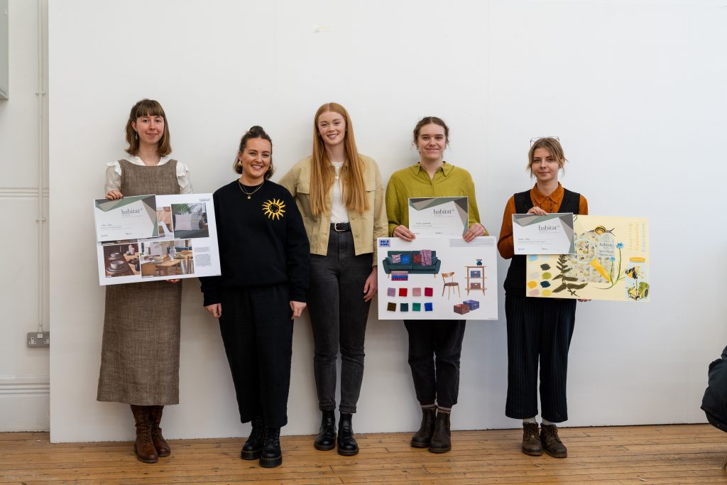 A photo of three Textile Design students and two Habitat designers smiling and addressing the camera. The Textile Design students are holding their portfolio boards and certificates from an awards ceremony. 