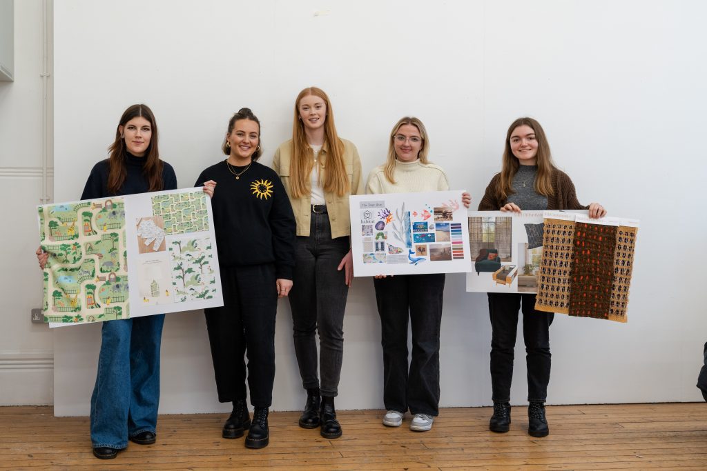 A photo of three Textile Design students and two Habitat designers smiling and addressing the camera. The Textile Design students are holding their portfolio boards.
