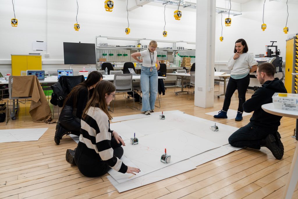 A photo in the Tech Studio with four students using small robots to draw onto large pieces of paper on the floor.