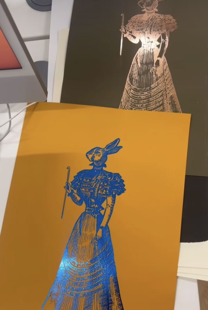 An orange sheet of paper with a blue foil print. The print is of a human body wearing a frilly dress but the head is taking the form of a rabbits head.  