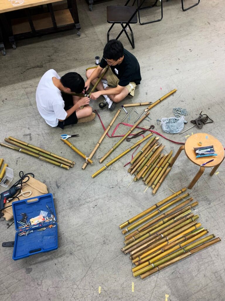 A photo of two students starting to assemble an art installation. The image is of two people sat on the floor working together to join bamboo pieces together. The floor is covered in bamboo, nails, rope and other various tools and materials. 