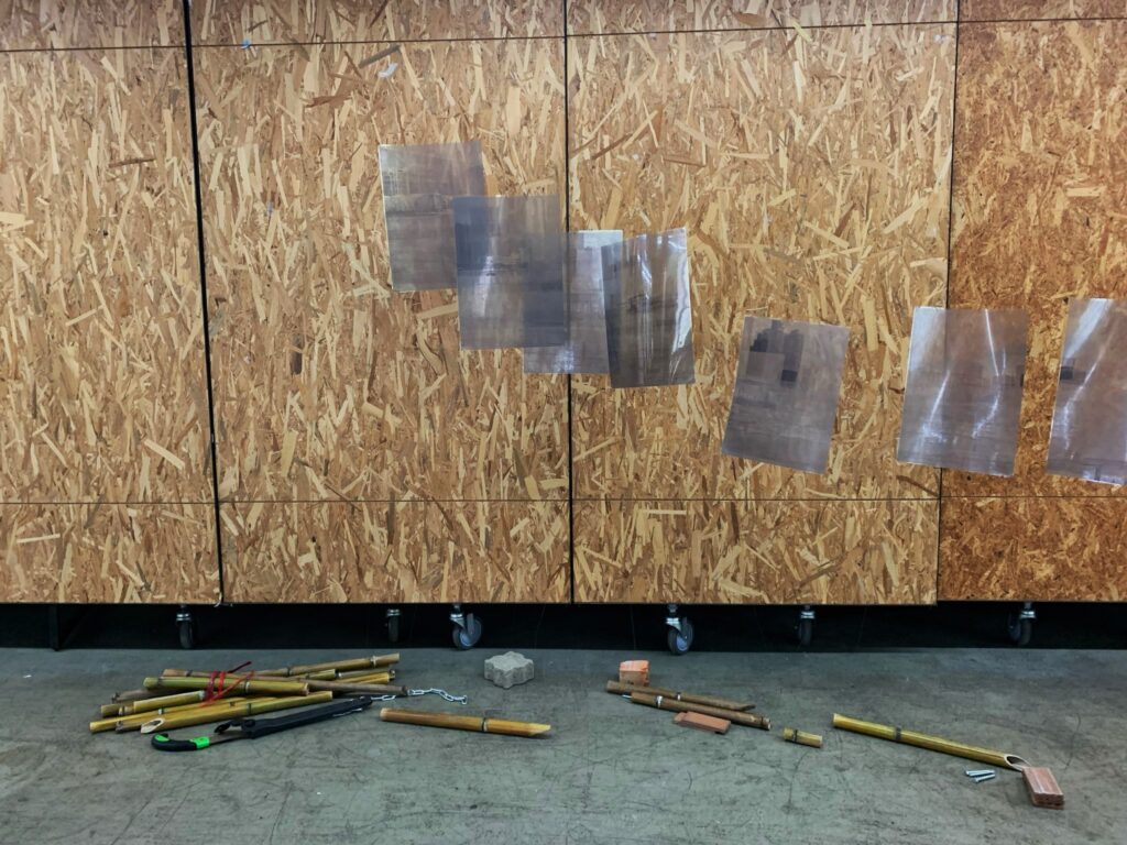 A photo of an art installation from the project: Urbanity from the Ocean, called TRANSFORMATIVE COOPERATION. 

The installation has various pieces of bamboo and brick scattered across the floor with handing pieces of plastic above them. 