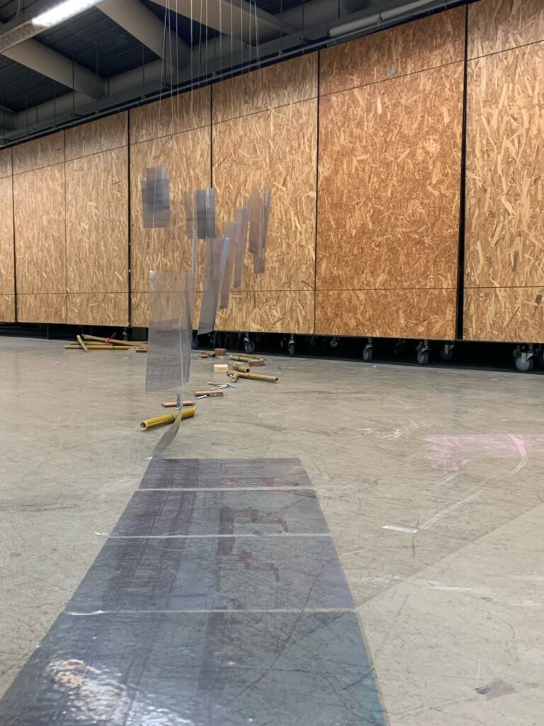 A photo of an art installation from the project: Urbanity from the Ocean, called TRANSFORMATIVE COOPERATION. 

The installation has various pieces of bamboo and brick scattered across the floor with handing pieces of plastic above them. 