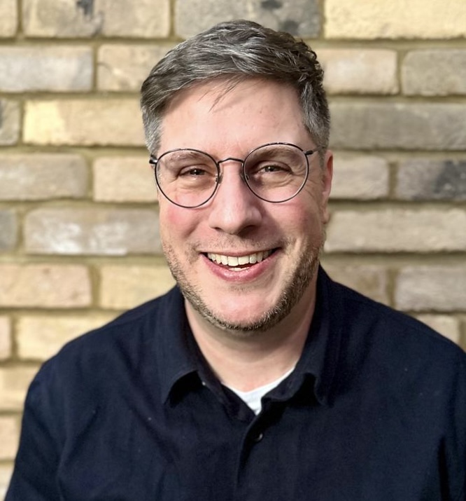 Photo of Ben Stopher. Ben has glasses and a navy shirt, in front of a brick wall.