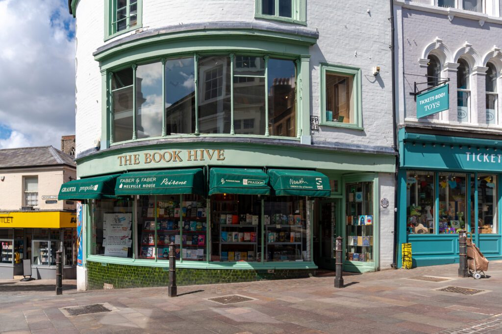 A photo of The Book Hive in Norwich. The image is of the shop front on a bright and sunny day. The shop is white brick with green windows. 