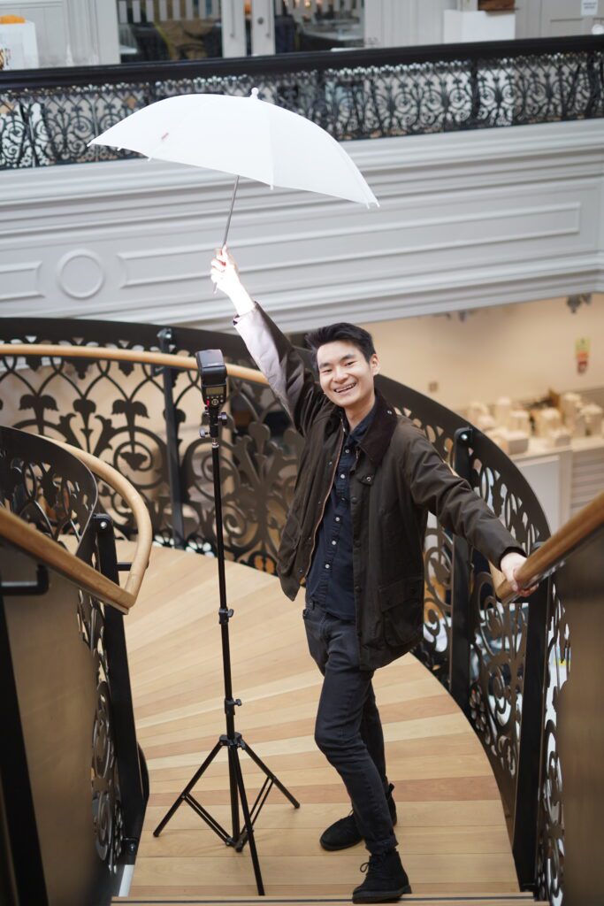 A photo of Desmond Lau in Boardman House, holding a lighting umbrella and smiling at the camera.