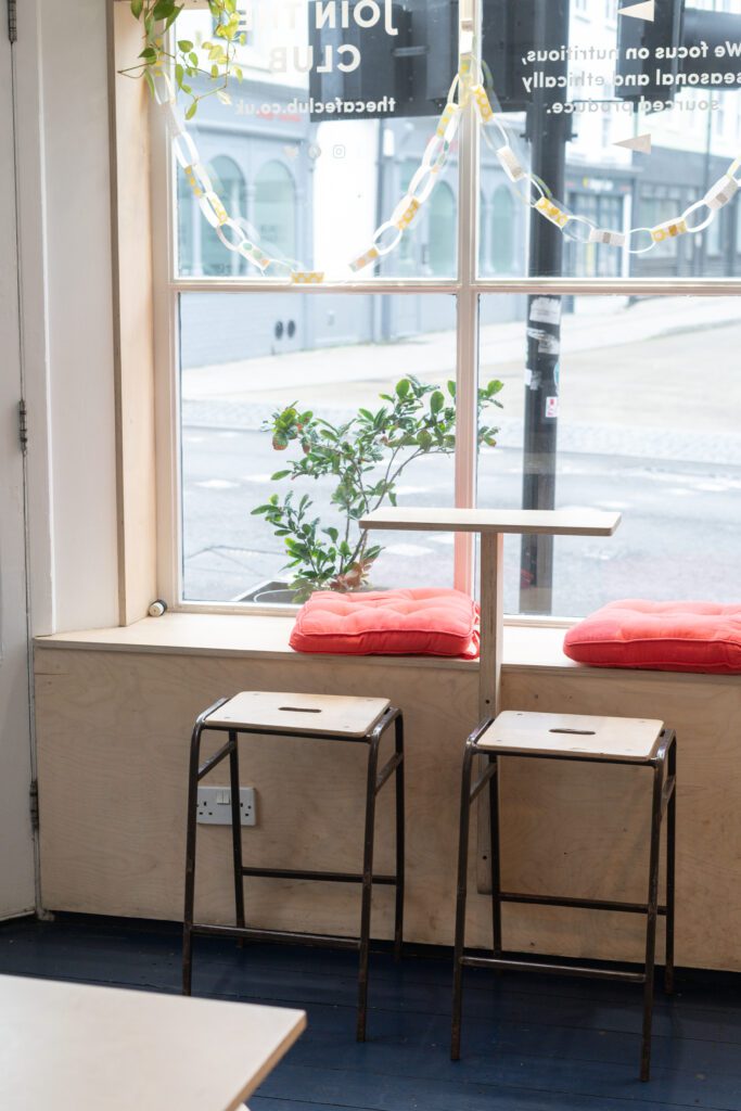 A photo of two wooden stools with red cushions and a tall table placed in front of a large window. 