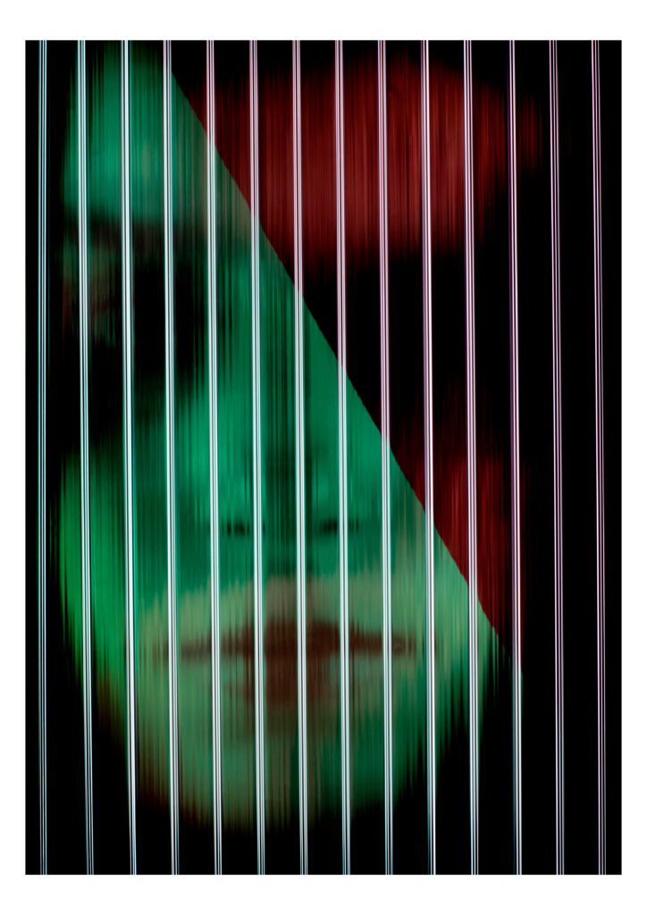 Photograph of a face with deep shadows and lit with a green light and a red stripe in the corner. There are vertical lines running across the image, distorting the face.