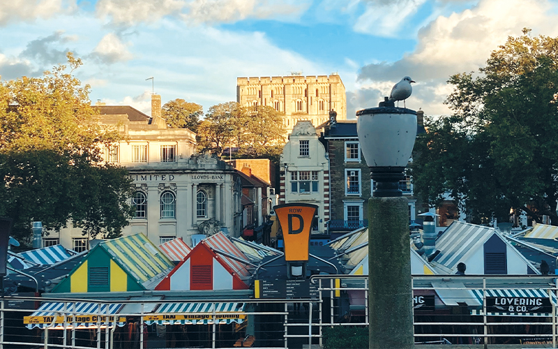 A photo of Norwich Market on a summers evening. The image is of the various different market stool roof tops that are all different colours. The Norwich Castle can be seen in the background of the photograph.
