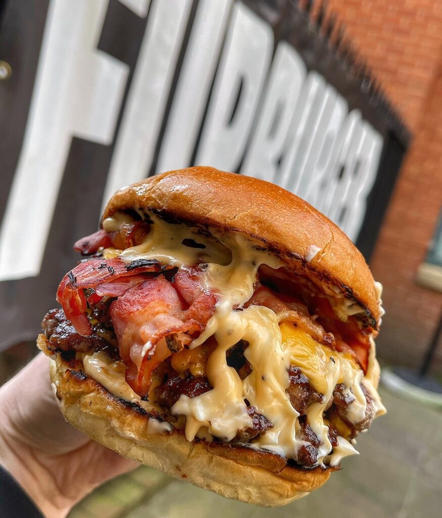 Close-up of a juicy burger held in a hand, featuring crispy bacon, melted cheese in a glossy, toasted bun. The background includes an out-of-focus banner and brick wall.