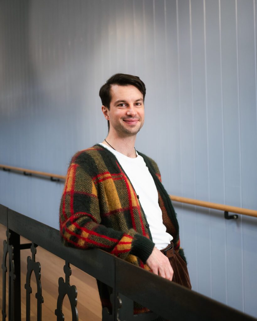 A person with short dark hair smiles while leaning on a black metal railing. They wear a white t-shirt under a colourful plaid cardigan. The background features a light blue panelled wall.