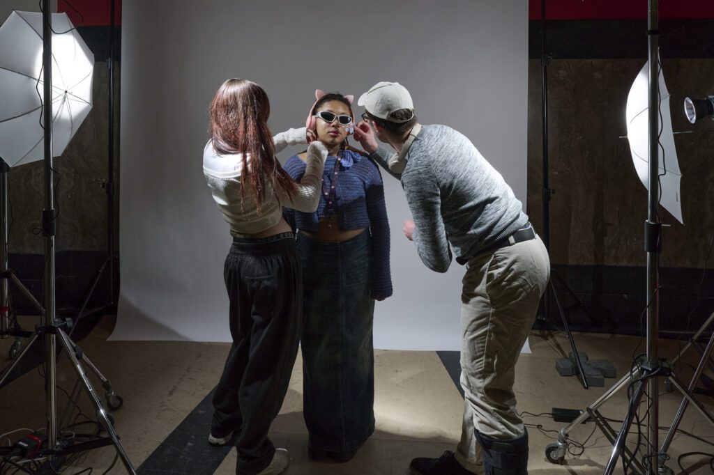 A person dressed in trendy clothing and sunglasses stands in a photo studio. Two individuals are adjusting their appearance. The studio setup includes a white backdrop, softbox lights, and equipment.