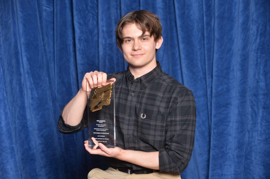 A person with short brown hair and a plaid shirt holds a clear award with gold accents against a blue curtain backdrop. They are smiling slightly and displaying the award prominently.