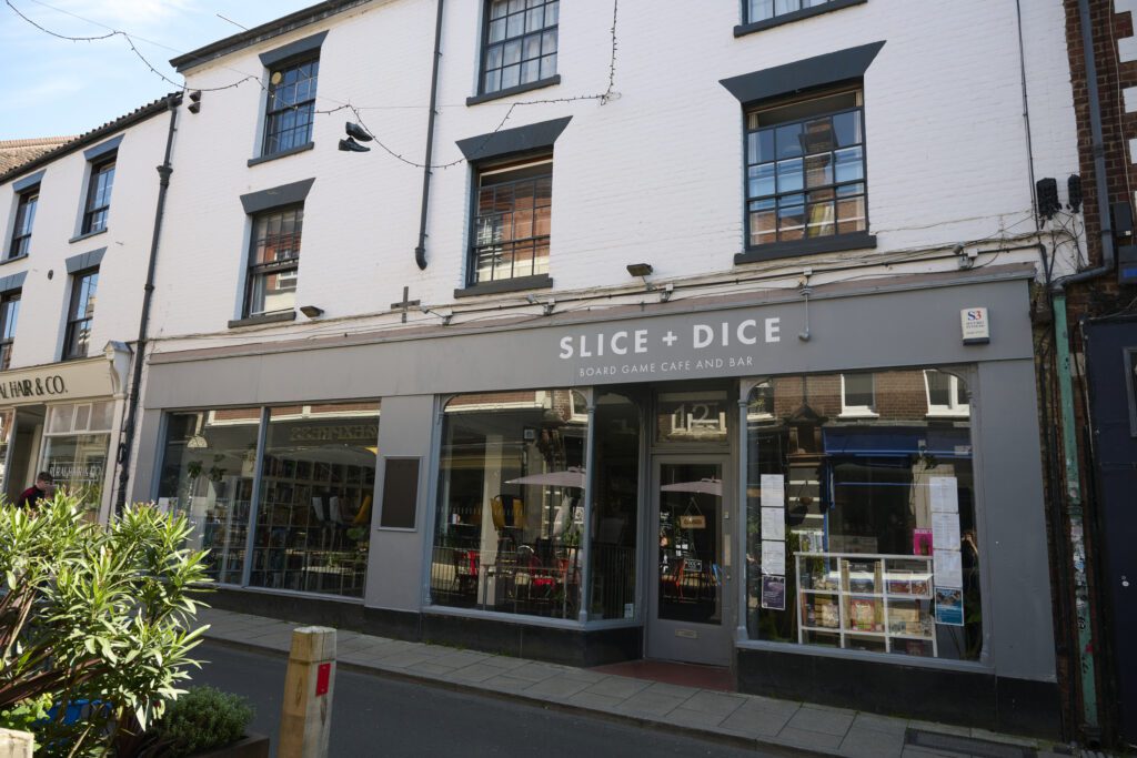 A street view of a white-painted building housing "Slice + Dice" board game cafe and bar. The storefront features large windows displaying various board games and posters.