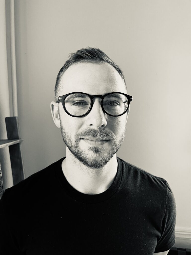 Black and white portrait of a person with short hair, wearing glasses and a black shirt, looking directly at the camera with a slight smile. The background is simple, possibly a wall, with part of a wooden structure visible.