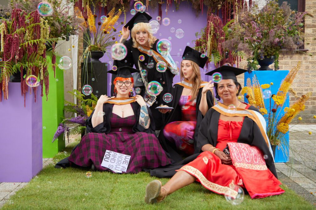 Four people wearing graduation gowns and caps celebrate outdoors. They are surrounded by colourful decorations and bubbles. There are bubbles floating in front of the group.