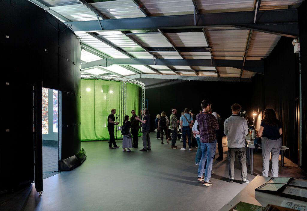 A group of people are gathered in an indoor space with a high ceiling. To the left, there's a green screen setup with lights. To the right, others are standing near tables with equipment. The room has a modern, industrial feel with metal beams and concrete floors.