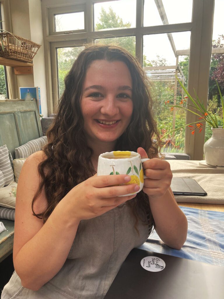 A woman with long hair smiles while holding a white mug with a yellow pattern. She is sitting in a cozy, sunlit room with large windows and various potted plants in the background.